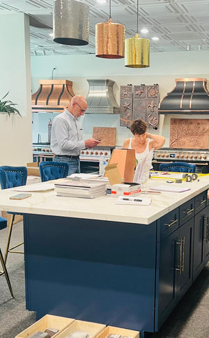 Two people in a kitchen showroom examining blue kitchen island and stainless steel range hood.